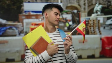 İspanyol bayrağı ve klasörleri taşıyan adam, şehrin bir caddesindeki inşaat sahasında düşünceli bir şekilde duruyor..