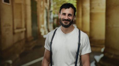 Sakallı İspanyol bir adam, üniversite kampüsünde kendine güvenen, tarihi bir mimariye sahip, günlük beyaz gömlek ve pantolon askısı giyen, akademik gelenek ve stil duygusu uyandıran,.