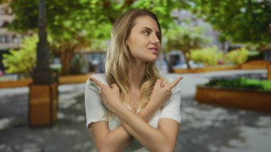 Parktaki bir kadın, yapraklı ağaçların altında dikilirken iki tarafı da çapraz kollarla işaret ederek doğal yeşil bir ortamda karar ya da yön öneriyor..