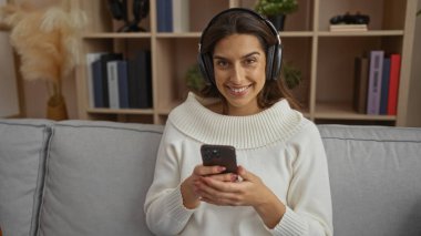 Kulaklık takan bir kadın, arka planda kitap rafları olan, rahat ve modern bir ev ortamını gösteren rahat bir oturma odasında telefon tutarken gülümsüyor..