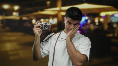 Gece caddesindeki restoranın terasında kameralı genç adam tefekkür ve tarzını ifade ediyor..