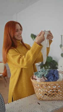 Evde rahat bir çamaşırhanede elbiselerini katlayan, kızıl saçlarını sergileyen ve sarı bir kazak giyen, yeni temizlenmiş çamaşır sepetleriyle çevrili bir kadın..