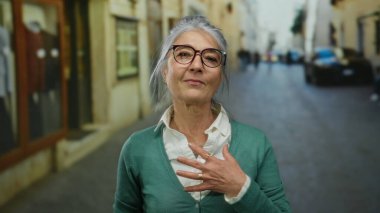 Gri saçlı ve gözlüklü yaşlı bir kadın şehir sokaklarında güvenle gülümsüyor, yeşil bir hırka giyiyor, şehirde sıcaklığını ve bilgeliğini iletiyor..