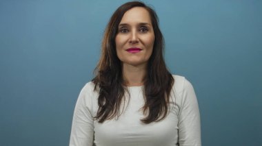 İzole edilmiş mavi bir arka plana sıcak bir şekilde gülümseyen bir kadın elleri sımsıkı bağlanmış, beyaz bir gömlek ve pembe bir ruj sürmüş, neşe ve dostluk duygusu iletiyor..