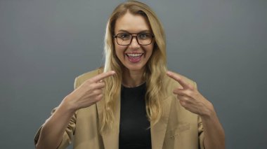 Gri arka planda, bej bir ceketle gülümseyen ve el kol hareketi yapan bir kadın..