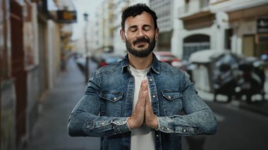 Sakallı İspanyol adam düşünceli bir şekilde şehir caddesinde duruyor, kot giyiyor, duacı hareketlerle duygularını ifade ediyor, ruhani bir doğa deneyimi sunuyor..