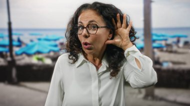 Deniz kenarındaki kıvırcık saçlı İspanyol kadın dikkatle dinliyor, şaşırıyor, plajda gözlük takıyor, merak ve ifadeyi vurguluyor..