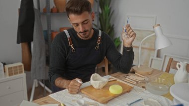 Sanatçının stüdyosundaki ahşap masada çiğ kil kâseyi fırça ve elle şekillendiren adam dikkatli bir konsantrasyon..
