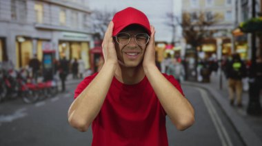 Kırmızı kurye üniforması giyerken trafiği bulanıklaştıran kalabalık bir sokak sahnesinde iki eliyle başını tutuyor..