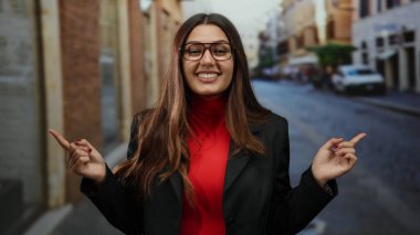 Sokakta gülümseyen bir kadın, iki eli gözlüklü ve kırmızı gömlekli, şehir ortamında, tercihini veya yönünü ima ediyor..