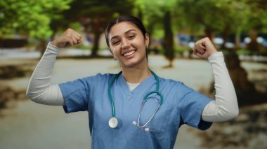 Önlüklü İspanyol kadın güneşli bir parkta esnek kollarıyla güven içinde poz veriyor. Açık hava sağlık hizmetlerinde kuvvet ve pozitifliği somutlaştırıyor..