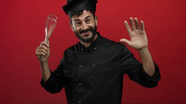 Sakallı İspanyol erkek aşçı neşeli bir şekilde çırpıcıyı canlı kırmızı arka planda tutuyor, aşçılık hevesini ve profesyonel kıyafetlerini sergiliyor..