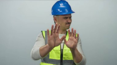 Yansıtıcı yelek ve şapka giyen İspanyol genç adam, tecrit edilmiş beyaz bir arka plana karşı reddedici bir jest yapıyor. Tereddüt veya ihtiyatı dile getiriyor..