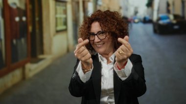 Orta yaşlı İspanyol bir kadın şehir sokaklarında parmaklarıyla kalp hareketi yapıyor, gülümsüyor, gözlük takıyor, siyah bir takım elbise giyiyor, mutluluğu ve arkadaşlığı somutlaştırıyor..