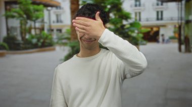 İnsan, şehir caddesinde gözleri kapamak için elini kullanıyor. Arkasında bulanık binalar ve yeşillikler var. Utanç..