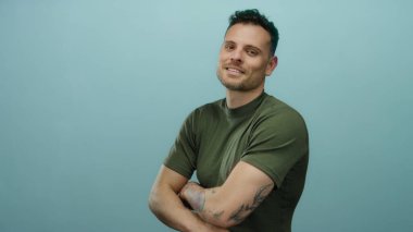 Dövmeleri ve çapraz kolları olan genç adam mavi bir arka plana karşı duruyor, yeşil bir gömlek giyiyor, modern saç stiliyle gülümsüyor, şık ve çekici bir ifade sergiliyor..
