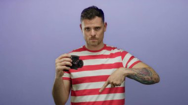 Dövmeli bir adam mor arka planda kamerayı tutuyor, aşağı bakıyor, çizgili gömlek giyiyor, kafa karışıklığı ya da merakını ifade ediyor..