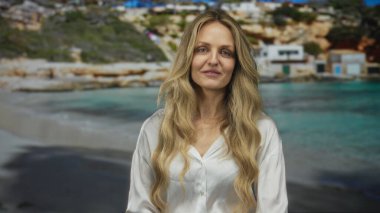 Sahilde gülümseyen beyaz tişörtlü genç sarışın kadın. Arka planda bulanık bir kıyı manzarası var. Huzurlu ve rahat bir açık hava anı yakalıyor..