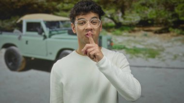 Ormandaki yeşil cipin yanında sessizlik için dudaklarına beyaz süveter ve gözlük takan bir adam..