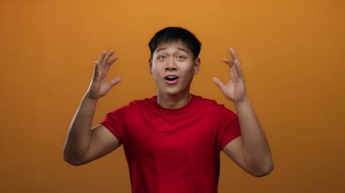 Duygularını izole edilmiş turuncu bir arka plana karşı ifade eden genç adam heyecan, kararlılık ve neşe vurguluyor, canlı kırmızı bir gömlek giyiyor ve güçlü jestler sergiliyor..