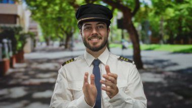 Sakallı genç adam pilot üniforması giyiyor güneşli bir sokakta el çırpıyor..