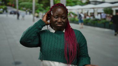 Pembe örgülü yeşil kazaklı bir kadın şehir caddesinde dikiliyor. İnsanlar bulanık arka planda yürürken belirsiz görünüyor..