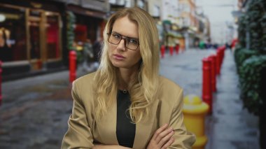 Sarışın ve gözlüklü bir kadın şehir caddesinde kollarını kavuşturarak dalgın dalgın dalgın ve şehir ortamında biraz üzgün görünüyor..