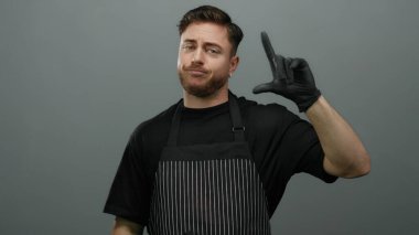 Önlüklü, sakallı genç adam gri duvarlı arka planda ezik işareti takıyor..