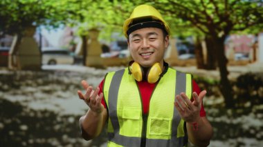 Emniyet yeleği ve kasket takan Asyalı genç adam ağaçlarla ve şehir manzarasıyla güvenle gülümsüyor..