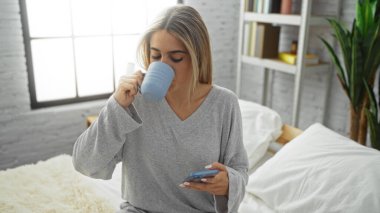 Kadın, rahat yatak odası ortamında telefona bakarken kahveden zevk alıyor. Rahatlama ve modern teknolojiyi harmanlayan huzurlu bir sabah rutinini vurguluyor..