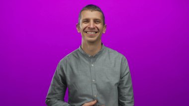 Canlı pembe arka planda gülümseyen bir adam, gözleri kapalı, neşeli bir ifade ve kapalı bir iç mekanda güçlü ışıklandırmayla günlük bir stil yakalıyor..