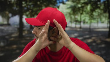 Kırmızı üniformalı teslimatçı, gün ışığında yeşil bir parkta başını eğip dikkatle dinlerken kulak arkası yapıyor..