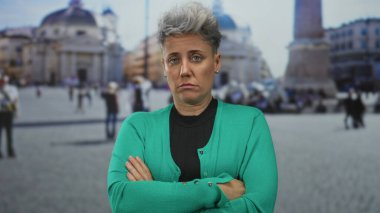 Kısa boylu, kaşlarını çatmış bir kadın açık gökyüzü altında süslü cephe ile büyük binanın dışında çaprazlama kollar;.