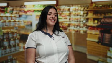 Kendine güveni tam genç bir kadın süpermarkette gülümsüyor. Etrafı ürün raflarıyla çevrili. Perakende satış ortamında kıvrımlı bir figür sergiliyor..
