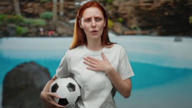 Kızıl saçlı bir kadın, otelde havuz başında futbol topu tutuyor, duygularını ifade ediyor, beyaz gömlek giyiyor, sakin bir yaz ortamı yaratıyor..