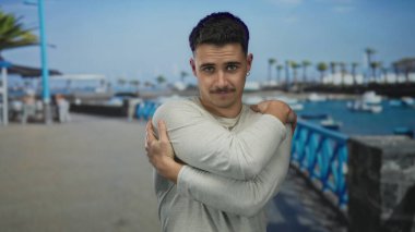 Genç İspanyol bir adam, sakin bir okyanus arkaplanı ve berrak mavi gökyüzü ile deniz kenarındaki bir gezinti alanında kendini kucaklıyor ve huzurlu bir açık hava ortamı sağlıyor..