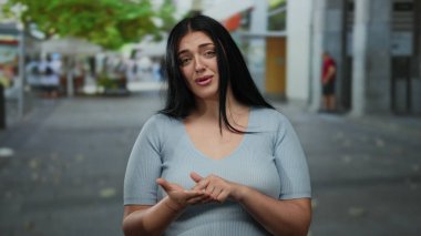 Bir şehir sokak ortamında yardım veya para için jest yapan bir kadın, samimi ve umut verici bir ifadeyle, kentsel bir çevre ile çevrili, halk etkileşimini gösteren.