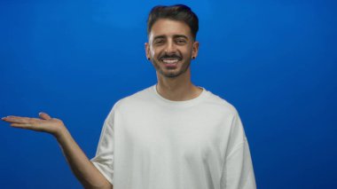 Beyaz gömlekli adam, stüdyoda gülümseyen avucunu uzatır ve mavi arka planda bir jest yapar..