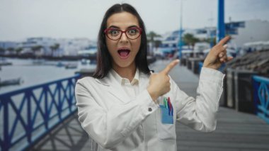 Üniformalı İspanyol kadın bilim adamı bulutlu gökyüzünün altında deniz kenarında gezinirken şaşırmış ve işaret etmiş..