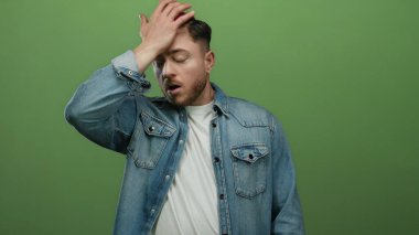 Kot ceketli genç adam yeşil arka planda duruyor. Şaşırmış görünüyor, duygularını ve kişiliğini izole edilmiş stüdyo sahnesinde aktarıyor..