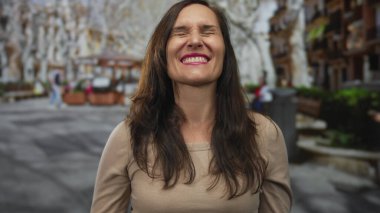 Canlı bir şehir caddesinde neşeli bir şekilde gülümseyen kadın, doğal güzelliği ve mutluluğu, bulanık mimari detaylarla çevrili, canlı bir açık hava ortamını yansıtıyor..