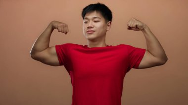 Genç Çinli bir adam kaslarını kırmızı bir gömlekle kahverengi bir arka plana yaslar ve gururlu, atletik bir duruşla gücünü ve güvenini gösterir..