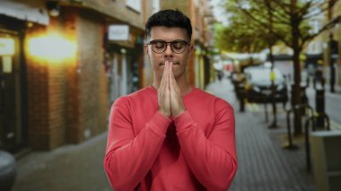 Kırmızı kazaklı, gözlüklü genç adam huzurlu bir şehir sokağında gün ışığında bulanık bir arka planda dua ediyor..