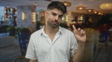 Sakallı bir adam, geniş bir otel binasında beyaz keten bir gömlek giyerken parmağını gösteriyor..