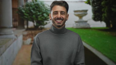 Genç adam yeşil yeşil yeşil parktaki taştan kolonun yakınındaki kameraya doğru gülümsüyor. Bulanık vazo ve yeşillik ile..