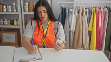 Yardım merkezinde gönüllü olarak çalışan bir kadın cep telefonu kullanıyor ve bağışlanan kıyafetlerle çevrili bir panoya yazıyor. Toplum çalışmalarını ve iç faaliyetleri ön plana çıkarıyor.