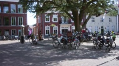 Büyüleyici bir Avrupa şehrinin kaldırım taşı meydanında, tuğlalı binaları ve dağınık manzarası olan büyük bir ağacın altına park edilmiş bisikletler..