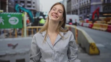 Yoğun caddede inşaat makineleriyle çerçevelenmiş, çizgili tişört giyen, gülümseyen sarışın genç bir kadın. Mutluluk..