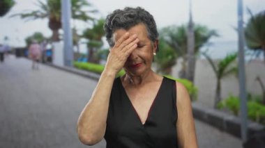 Yaşlı bir kadın, tropikal bitkiler ve plaj manzarasıyla çevrili deniz kenarında yürürken kendini iyi hissetmiyor..