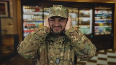 Kamuflaj üniformalı adam binadaki yankılanan gürültüde kulakları ve buruşukları kapatıyor..